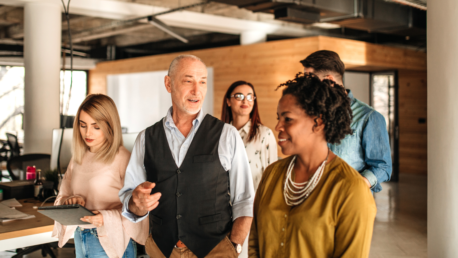 Diverse business team collaborating in a modern office, featuring an older male leader discussing with colleagues and a woman using a tablet.