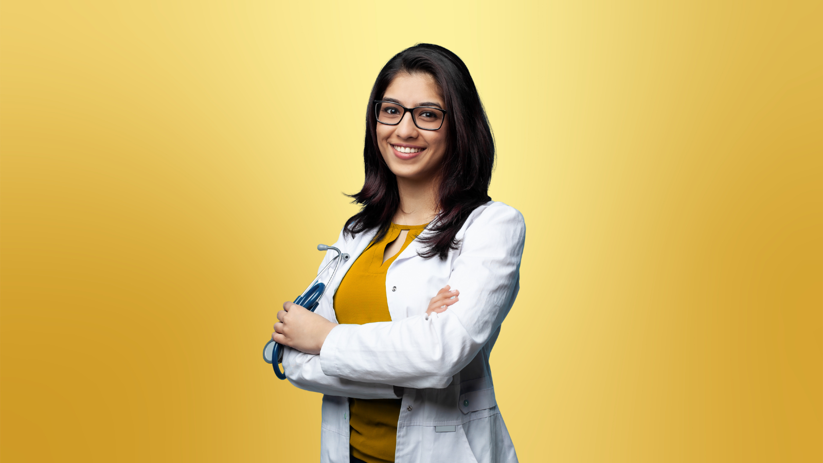 Smiling Indian woman doctor wearing glasses and a white lab coat, holding a blue stethoscope, standing confidently against a yellow background.