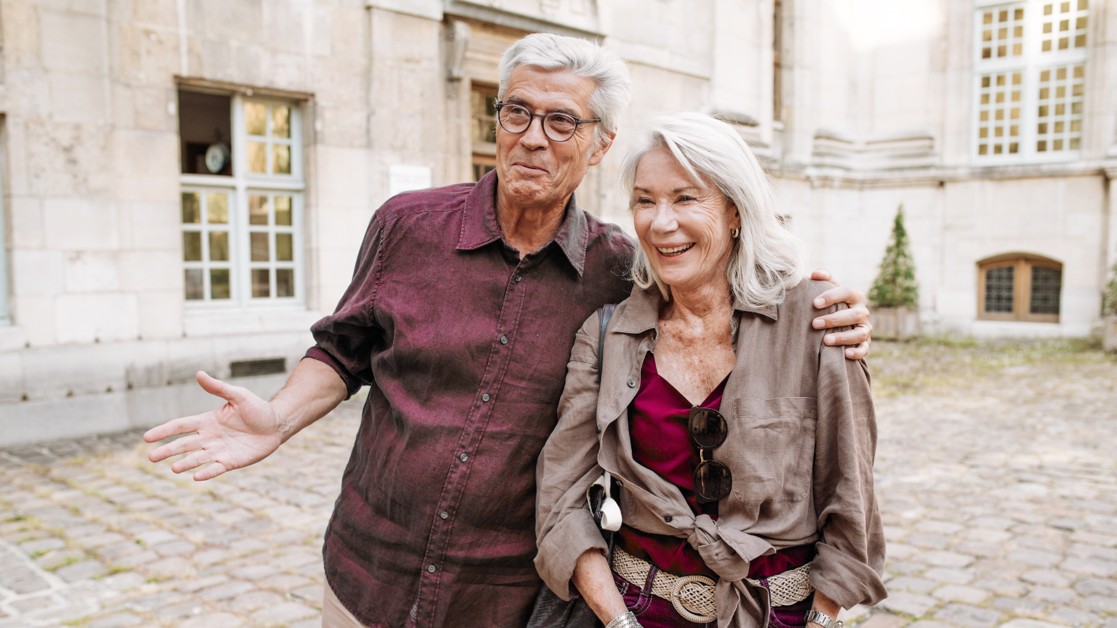 Happy senior couple with grey hair smiling and embracing on a cobblestone street in front of a historic stone building, enjoying their travel.