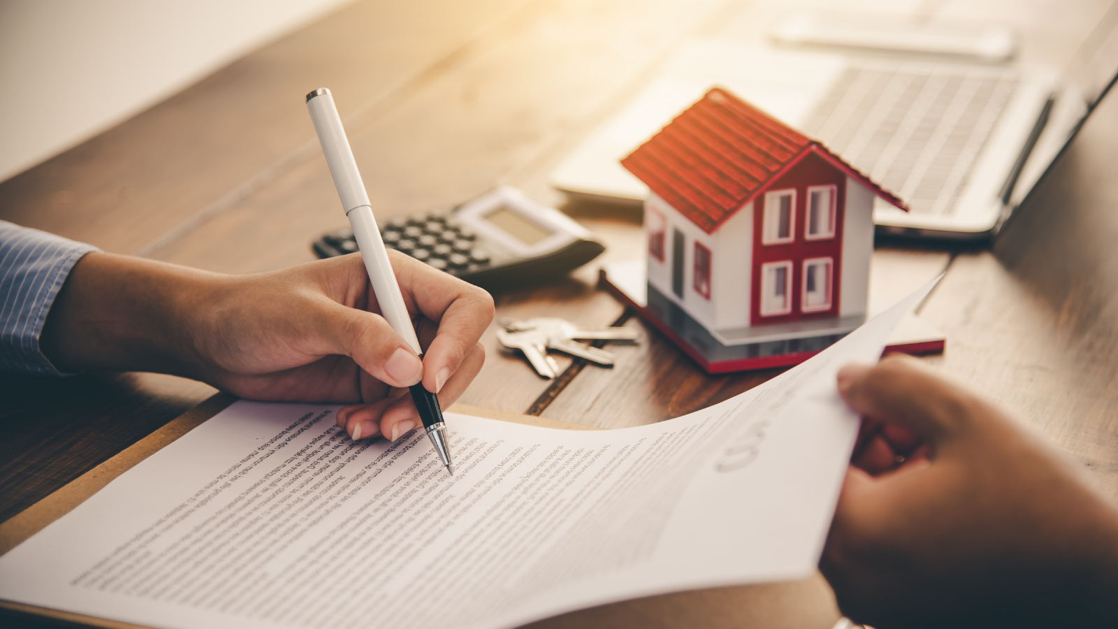 Person signing real estate contract with pen, model house and house keys nearby. Represents home purchase agreement or mortgage signing.